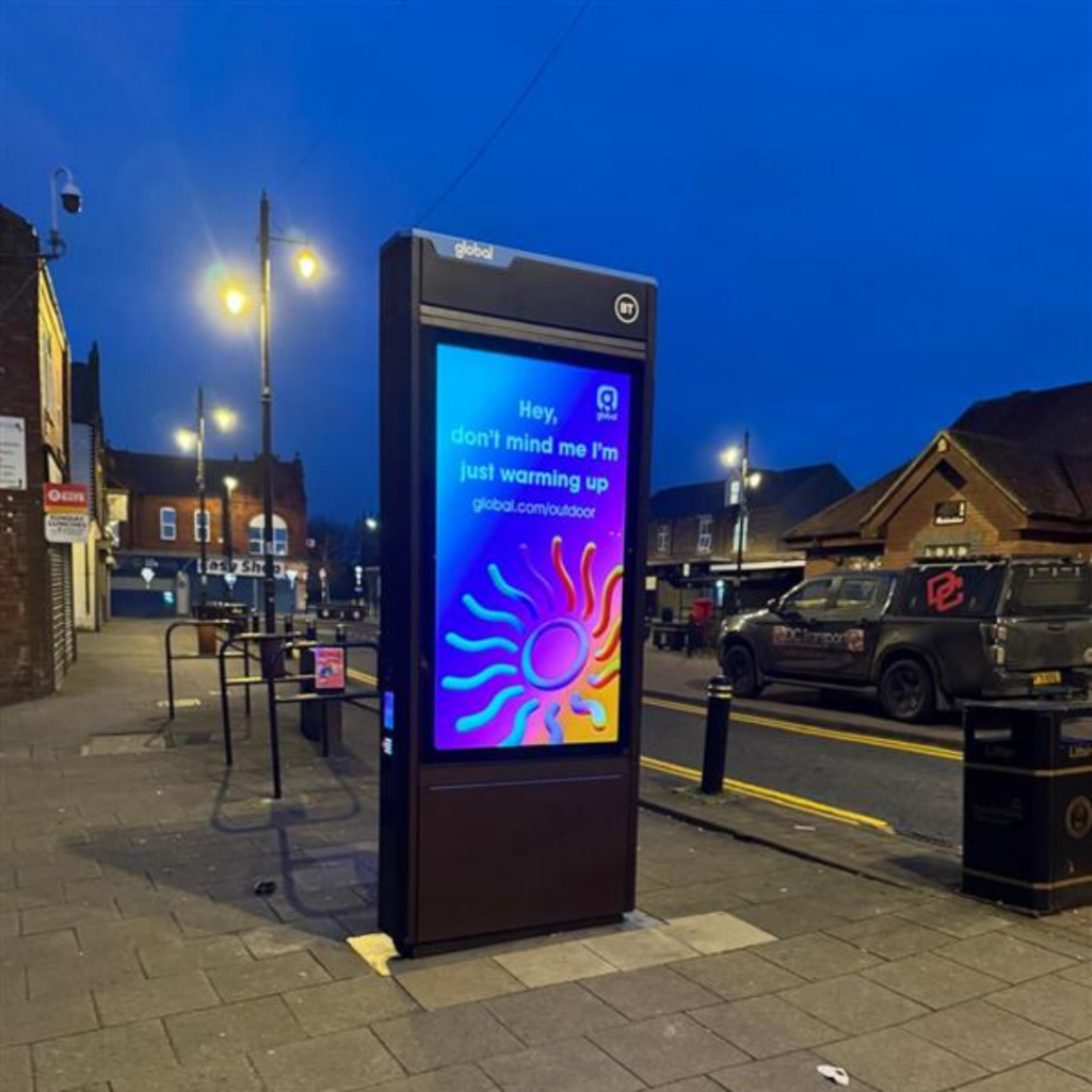 Illuminated freestanding digital advertising totem on a high street pavement at dusk, with a DC Transport and Engineering branded pickup truck parked in the background.