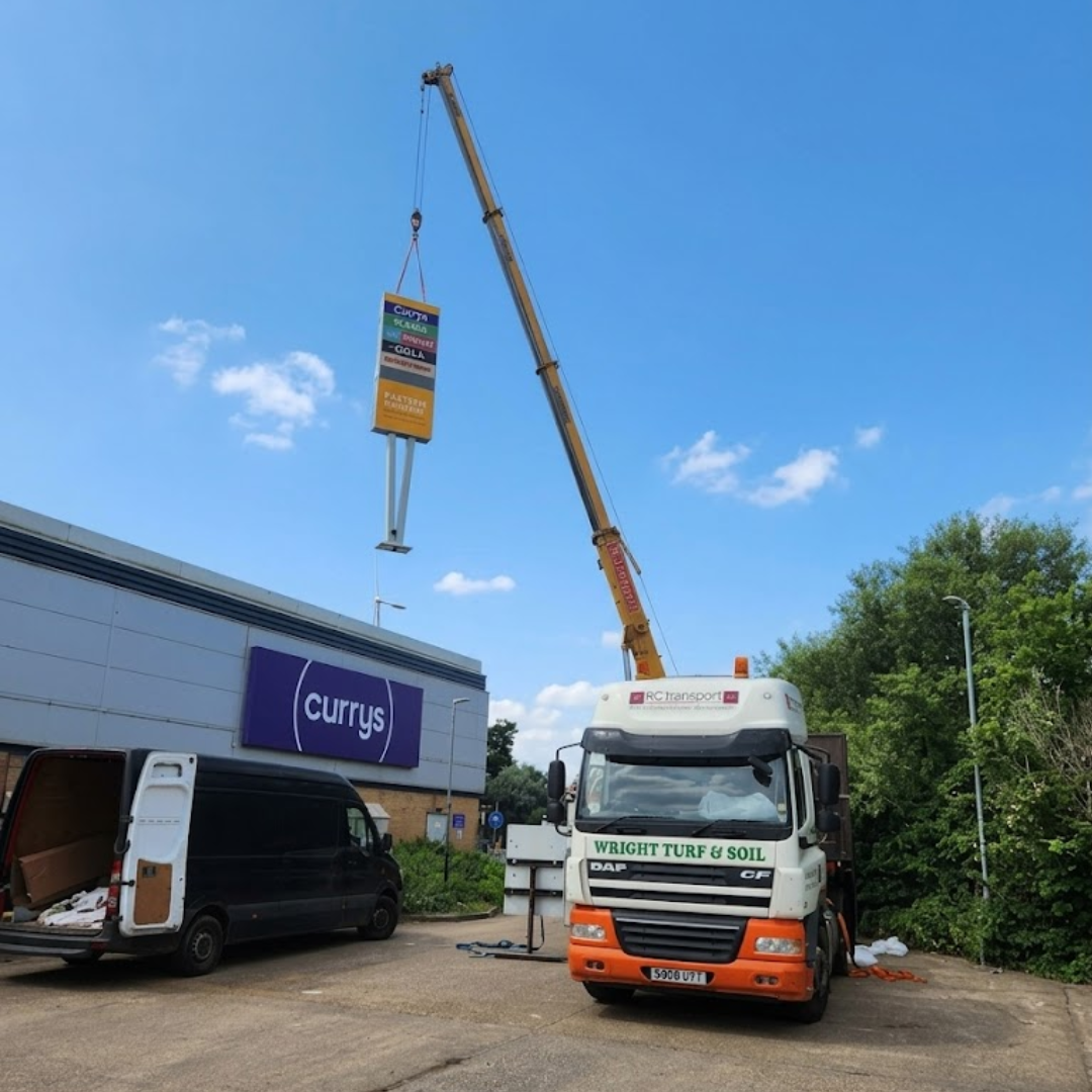 A heavy lifting crane hoisting a large retail park sign above a branded DC Transport commercial vehicle.